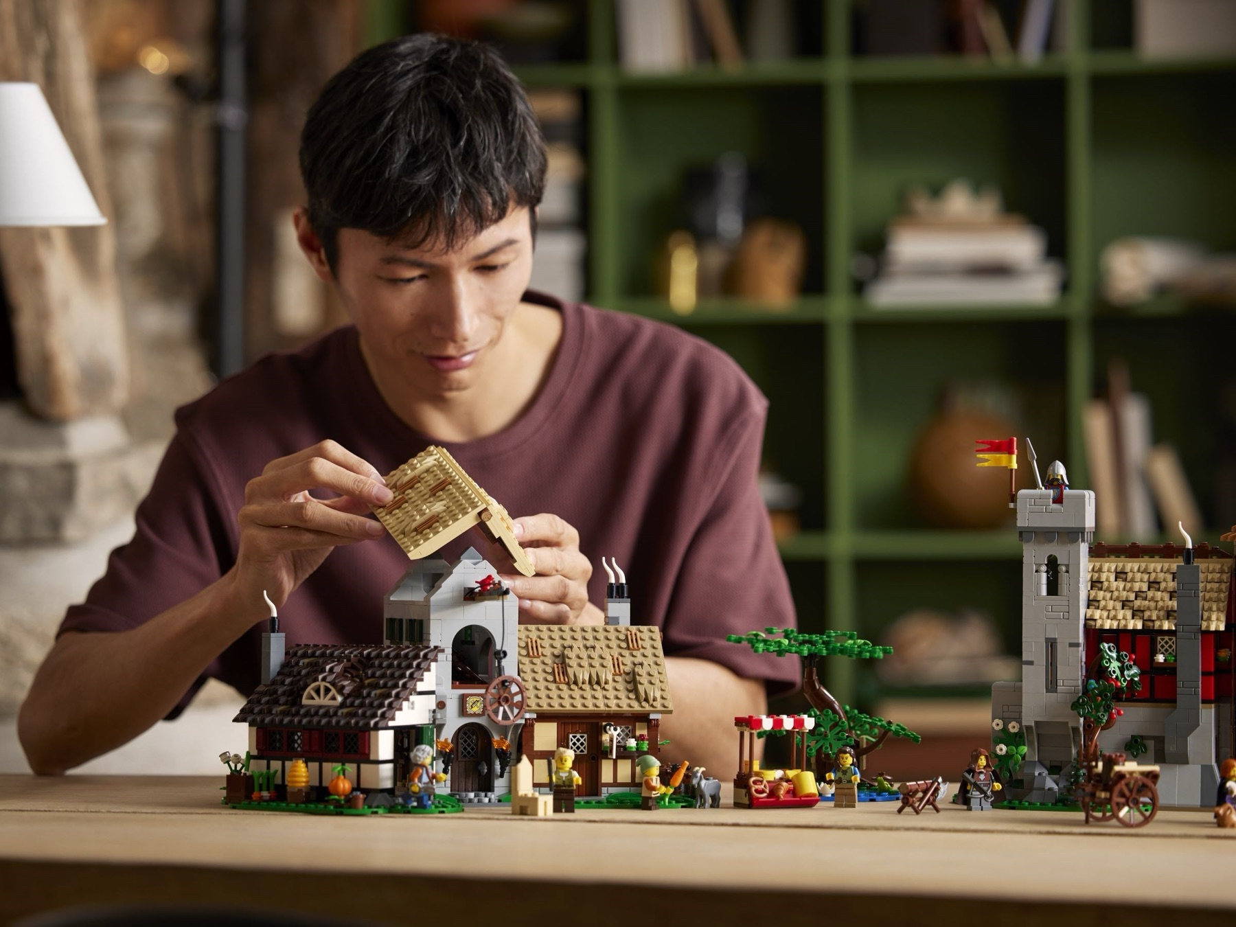Builder assembling LEGO Medieval Town Square 10332 with both village buildings, market stall and watchtower arranged across a table